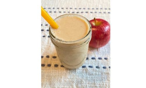 Apple pie smoothie in a small mason jar with a yellow straw and an apple displayed in the background.
